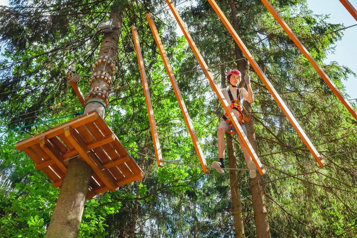 Teenager boy wearing safety harness passing hanging rope bridge obstacle at a ropes course in outdoor treetop adventure park